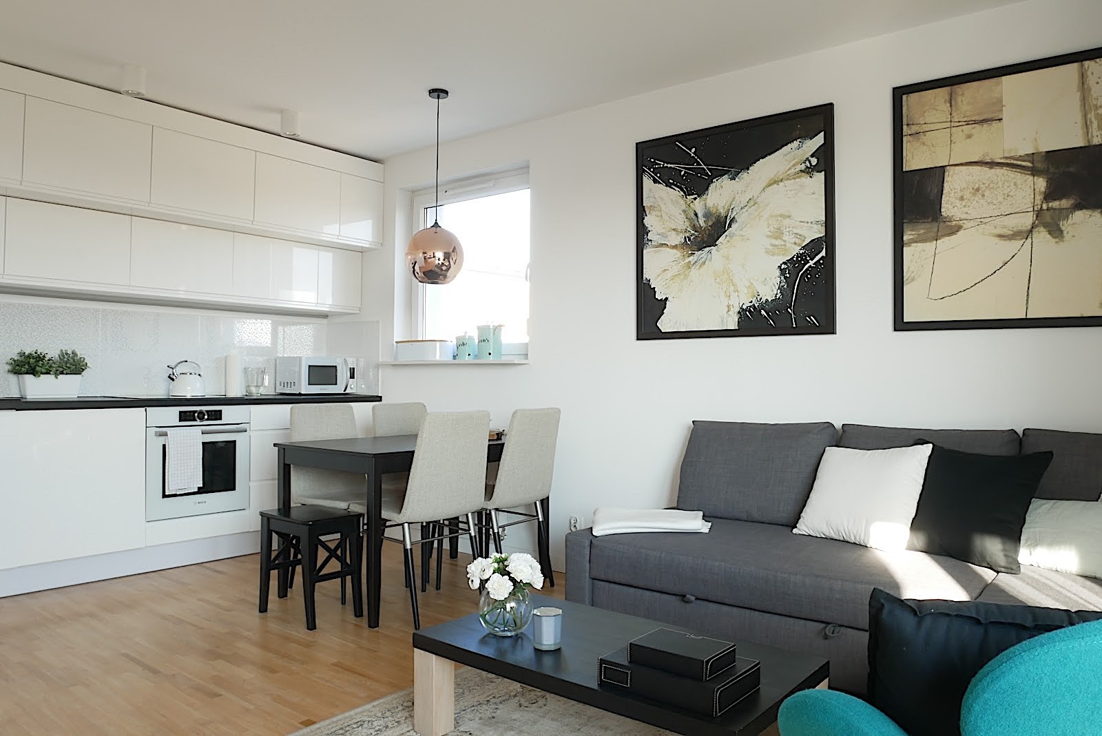 Jasne wnętrze mieszkania z aneksem kuchennym, ciemnym stołem i szarą sofą, ozdobione obrazami i miedzianą lampą wiszącą.