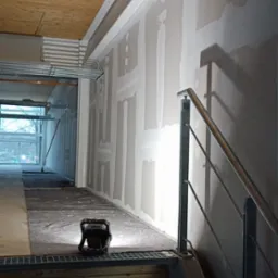 Ściana w trakcie wygładzania gipsem, widoczne łączenia płyt kartonowo-gipsowych, metalowe schody z kratą, lampa budowlana oświetlająca fragment podłogi zabezpieczonej materiałem.