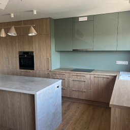 Meta-morfozy - Nowoczesna kuchnia z drewnianymi frontami szafek, wyspą kuchenną z blatem imitującym beton, zielonymi szafkami wiszącymi i designerską lampą wiszącą z czterema kloszami.