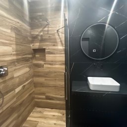 Meta-morfozy - Nowoczesna łazienka z prysznicem wykończonym drewnopodobnymi płytkami i umywalką na czarnym blacie z marmurowym wzorem, okrągłe lustro z podświetleniem.