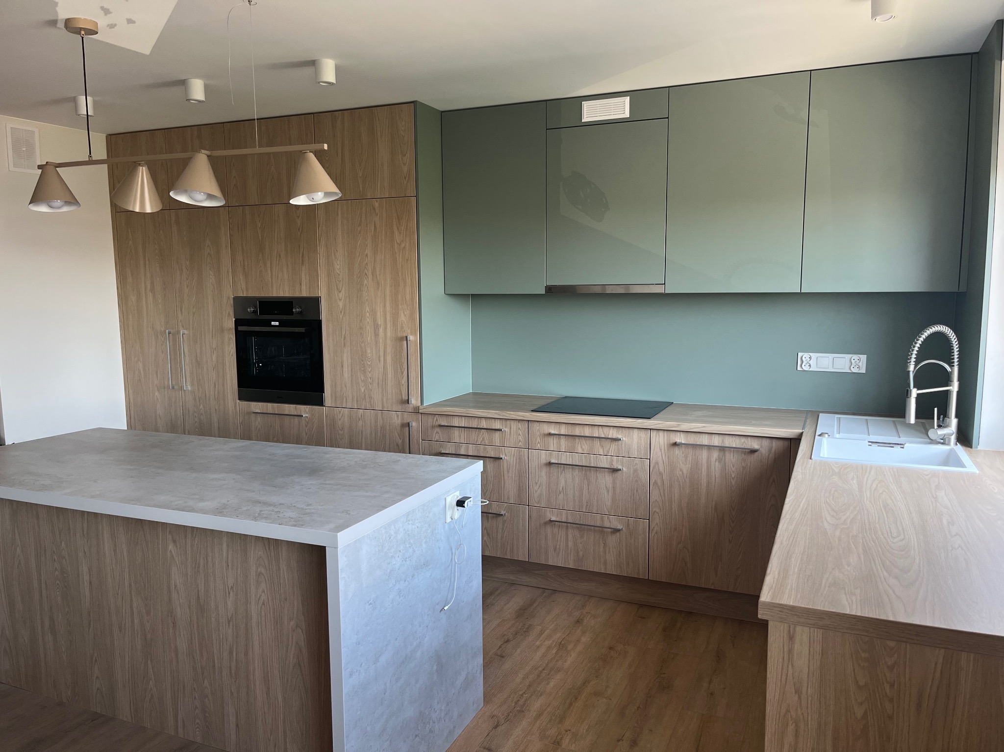 Nowoczesna kuchnia z drewnianymi frontami szafek, wyspą kuchenną z blatem imitującym beton, zielonymi szafkami wiszącymi i designerską lampą wiszącą z czterema kloszami.