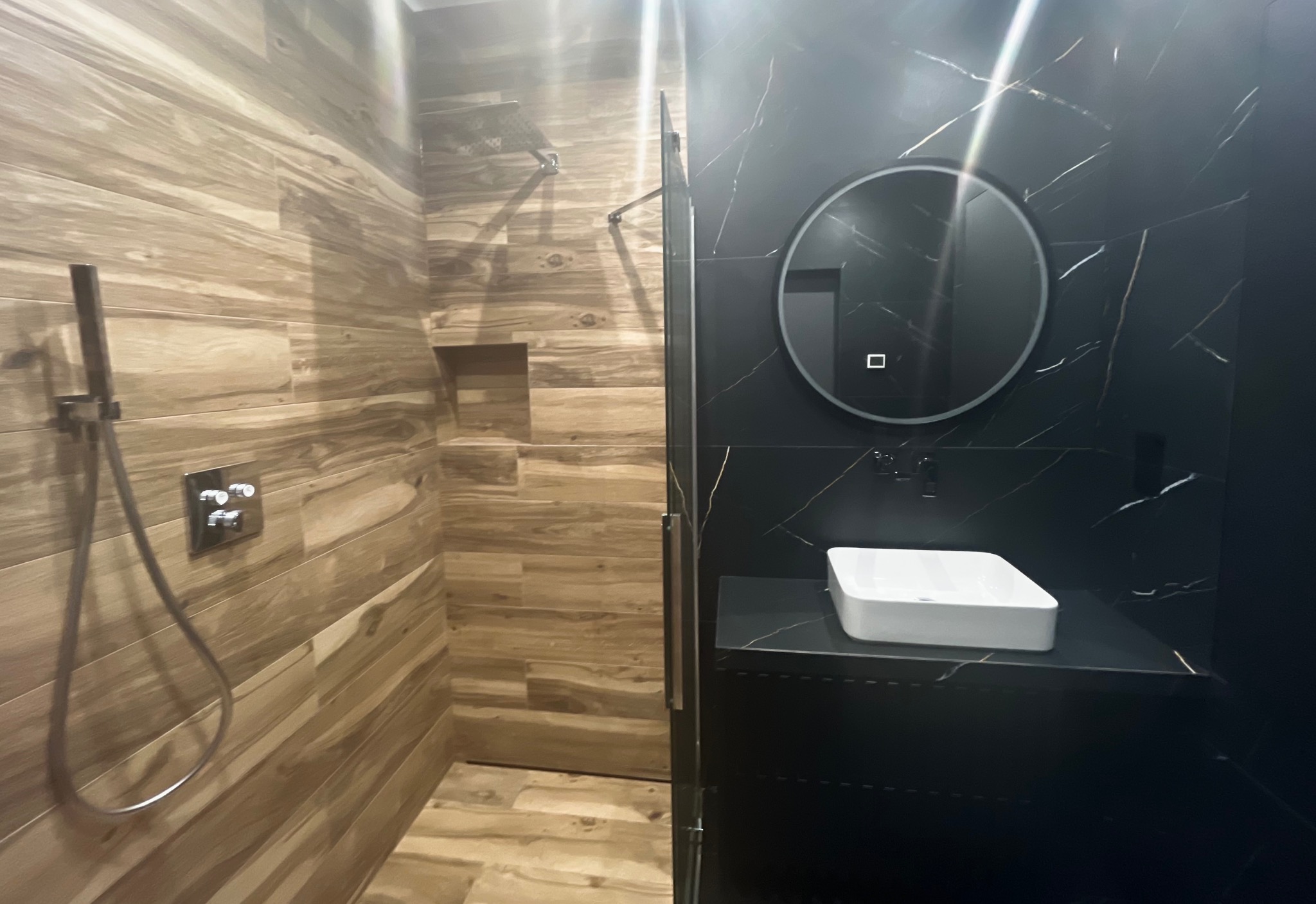 Nowoczesna łazienka z prysznicem wykończonym drewnopodobnymi płytkami i umywalką na czarnym blacie z marmurowym wzorem, okrągłe lustro z podświetleniem.