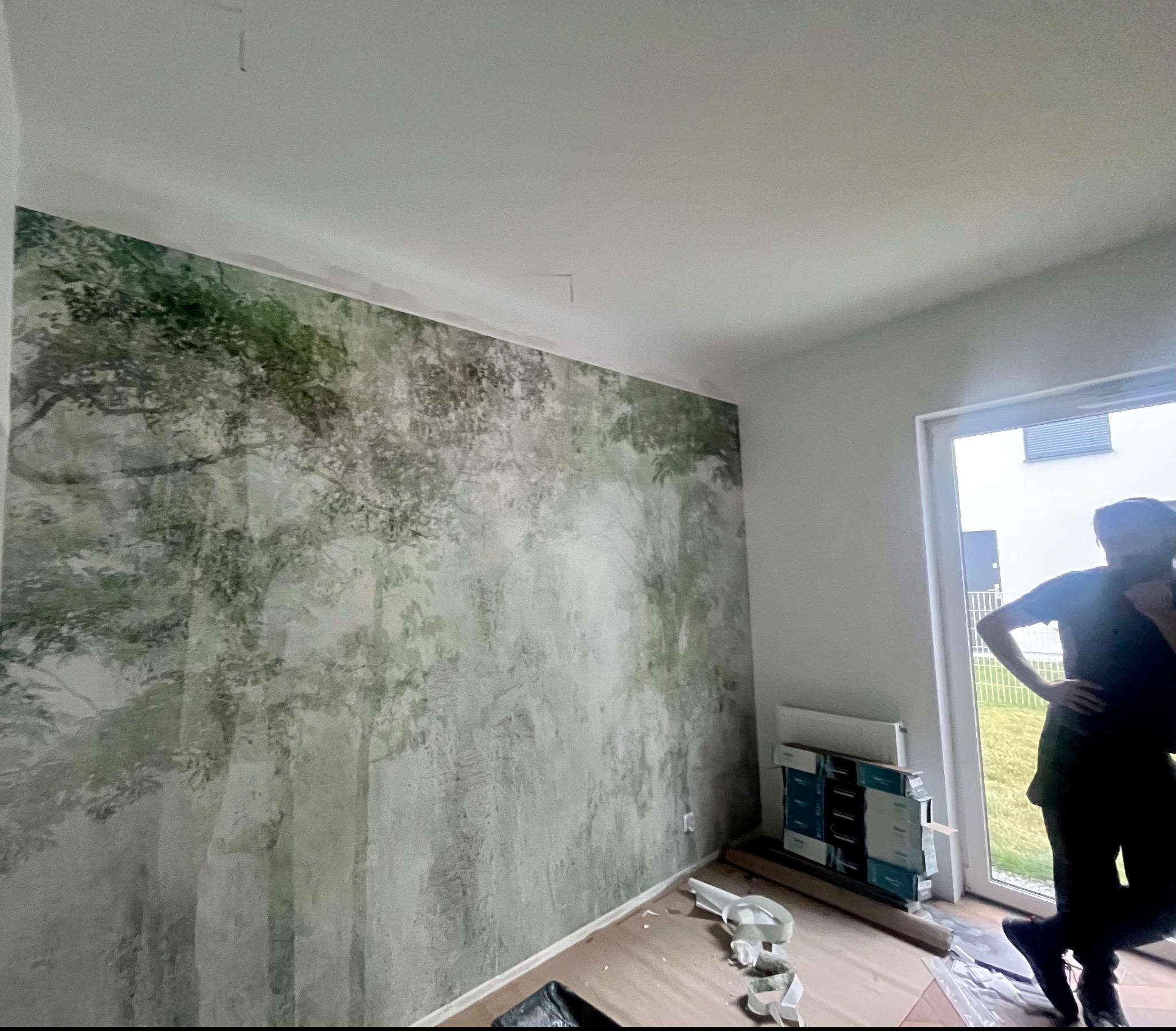 Ściana w pokoju z fototapetą przedstawiającą las w odcieniach zieleni i szarości, osoba w tle przy oknie, widoczne resztki po montażu tapety i opakowania po kleju.