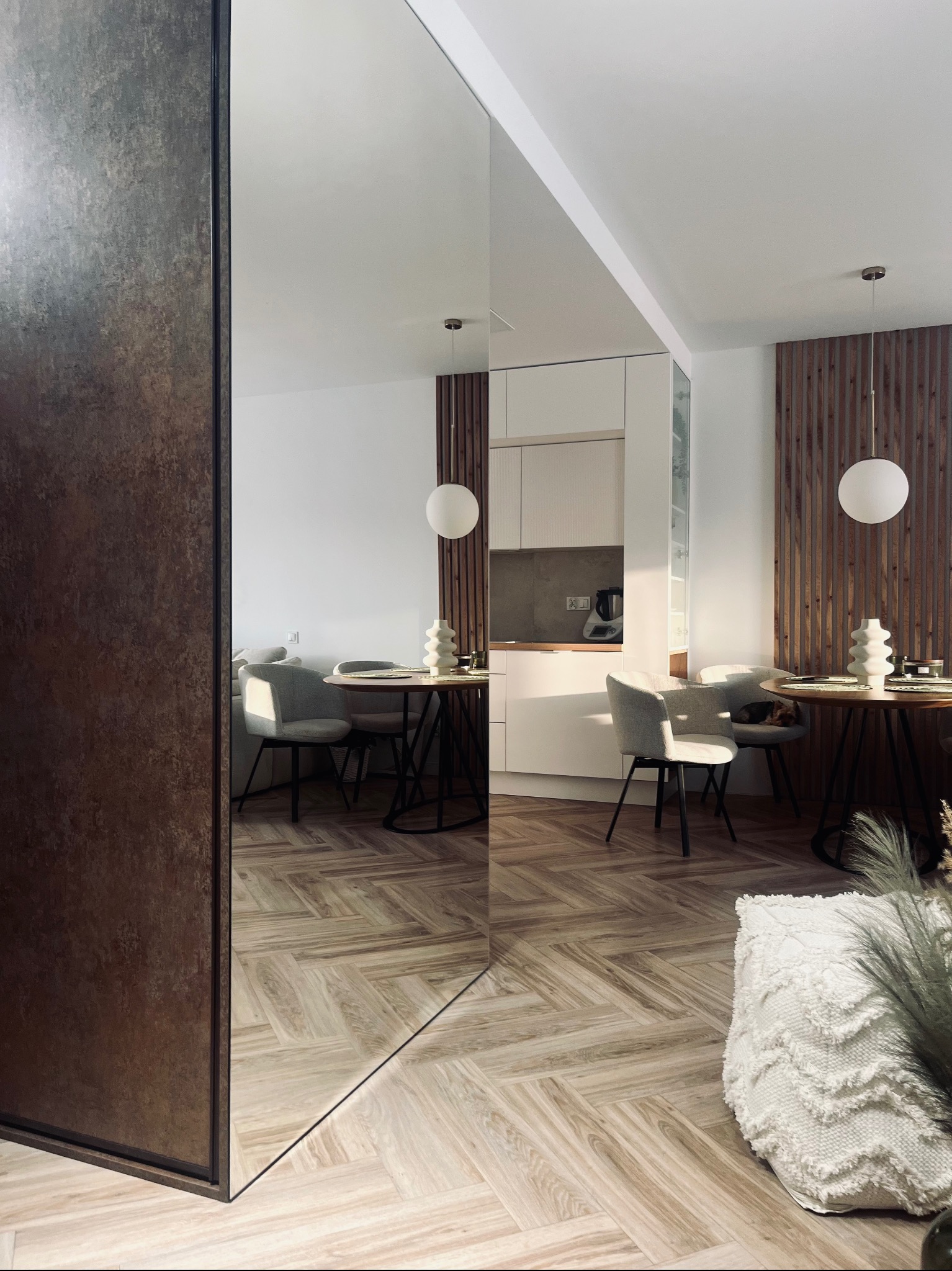 Nowoczesne wnętrze mieszkania z lustrzaną ścianą, parkietem w jodełkę, okrągłym stołem i drewnianą ścianką działową, oświetlone kulistymi lampami wiszącymi.