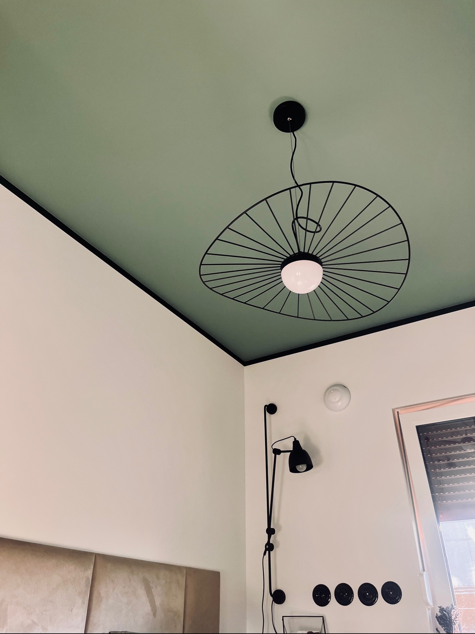 Nowoczesna sypialnia z zielonym sufitem i designerską lampą wiszącą z czarnymi prętami. Na ścianie kinkiet w stylu industrialnym i rząd czarnych gniazdek.