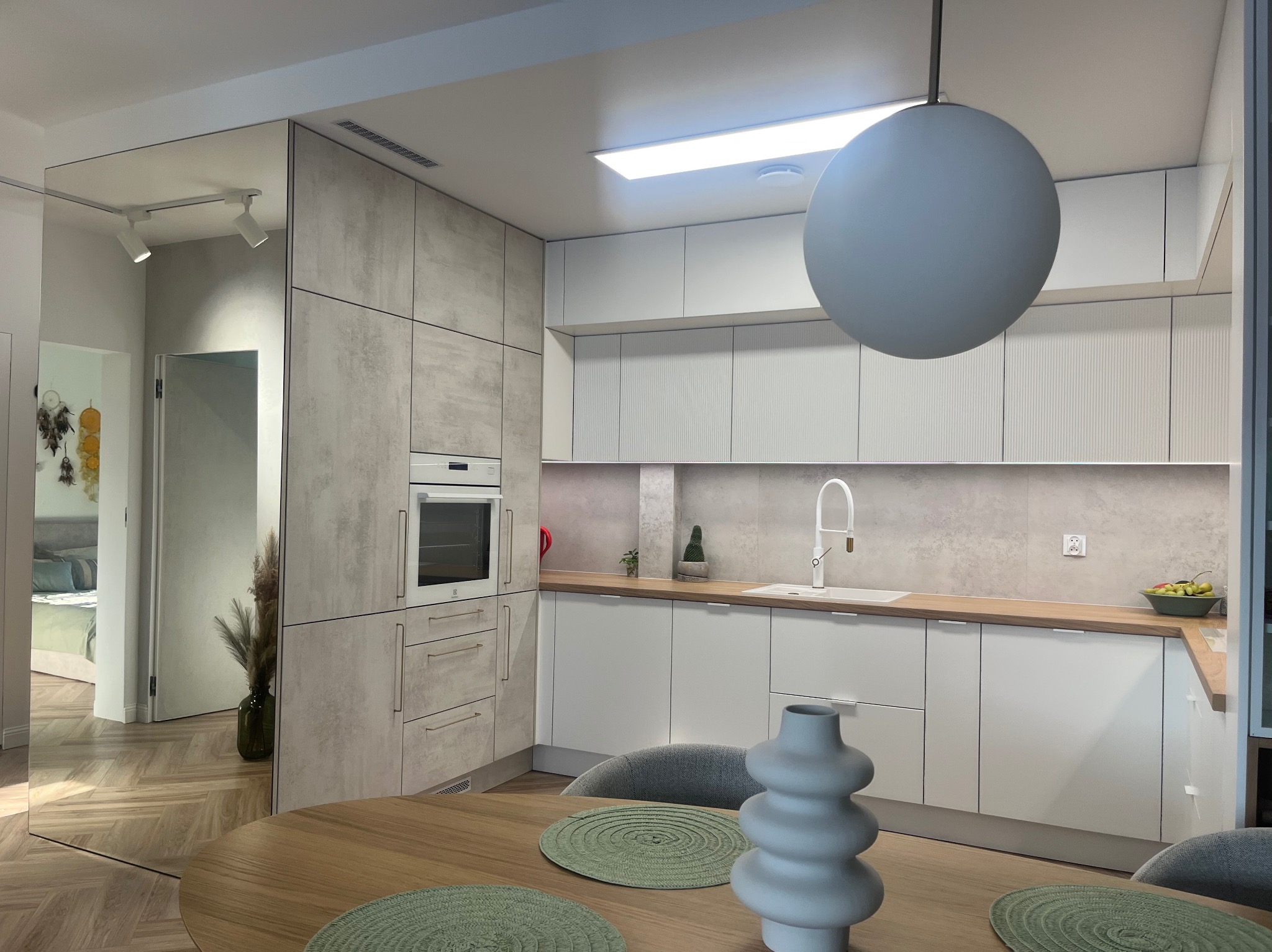 Nowoczesna kuchnia z elementami drewna, okrągły stół z dekoracyjnymi podkładkami i wazonem, widok na sypialnię z lustrzaną ścianą.