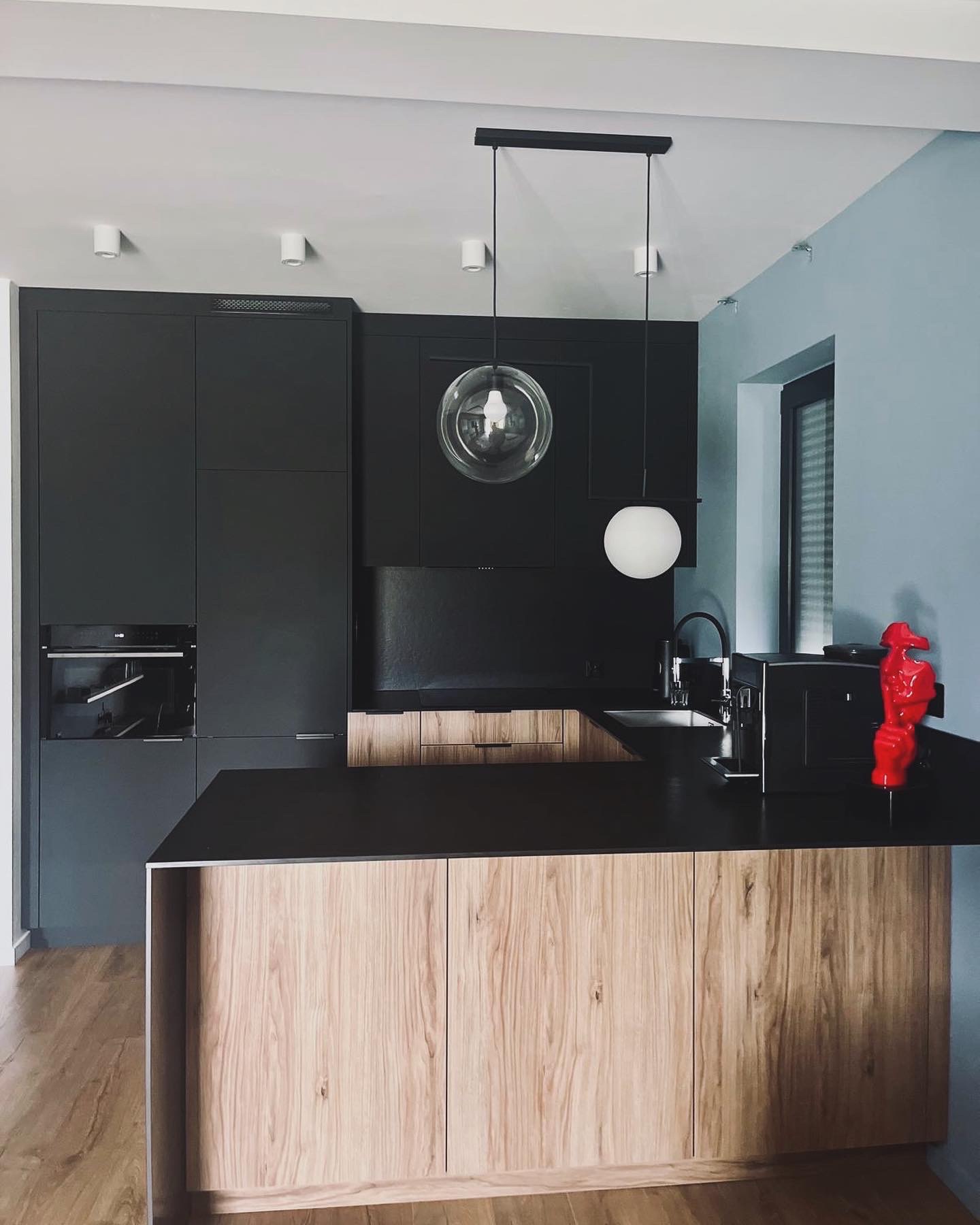 Nowoczesna kuchnia w odcieniach szarości i drewna z czarnym blatem, wiszącymi lampami kulami i czerwoną figurką na blacie