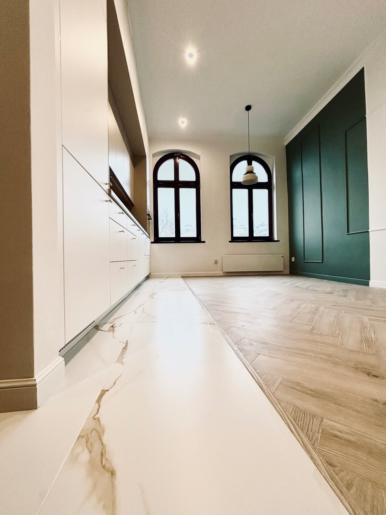 Nowoczesne wnętrze: biała zabudowa kuchenna z delikatnymi złotymi uchwytami, marmurowa podłoga przechodząca w parkiet ułożony w jodełkę, wysokie okna łukowe z czarnymi ramami, ściana w kolorze...