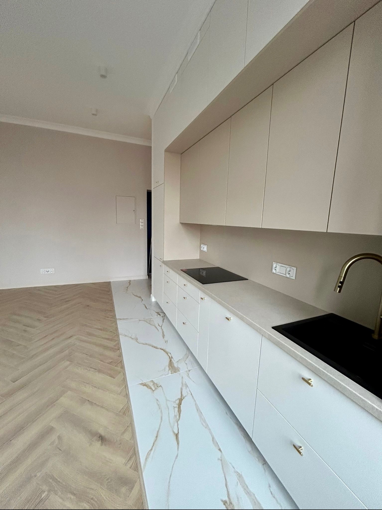 Nowoczesna kuchnia z białymi szafkami, złotymi uchwytami i marmurową podłogą przechodzącą w parkiet ułożony w jodełkę.