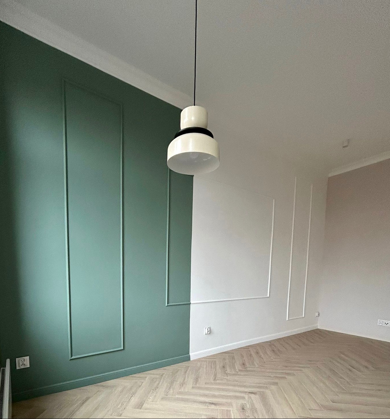 Pomalowane na zielono i biało ściany z dekoracyjnymi listwami, parkiet ułożony w jodełkę, wisząca lampa w stylu retro.