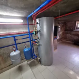 Szary zasobnik ciepłej wody użytkowej z niebieskimi i czerwonymi rurami instalacji grzewczej w piwnicy z surowym betonowym sufitem i płytkami na podłodze.