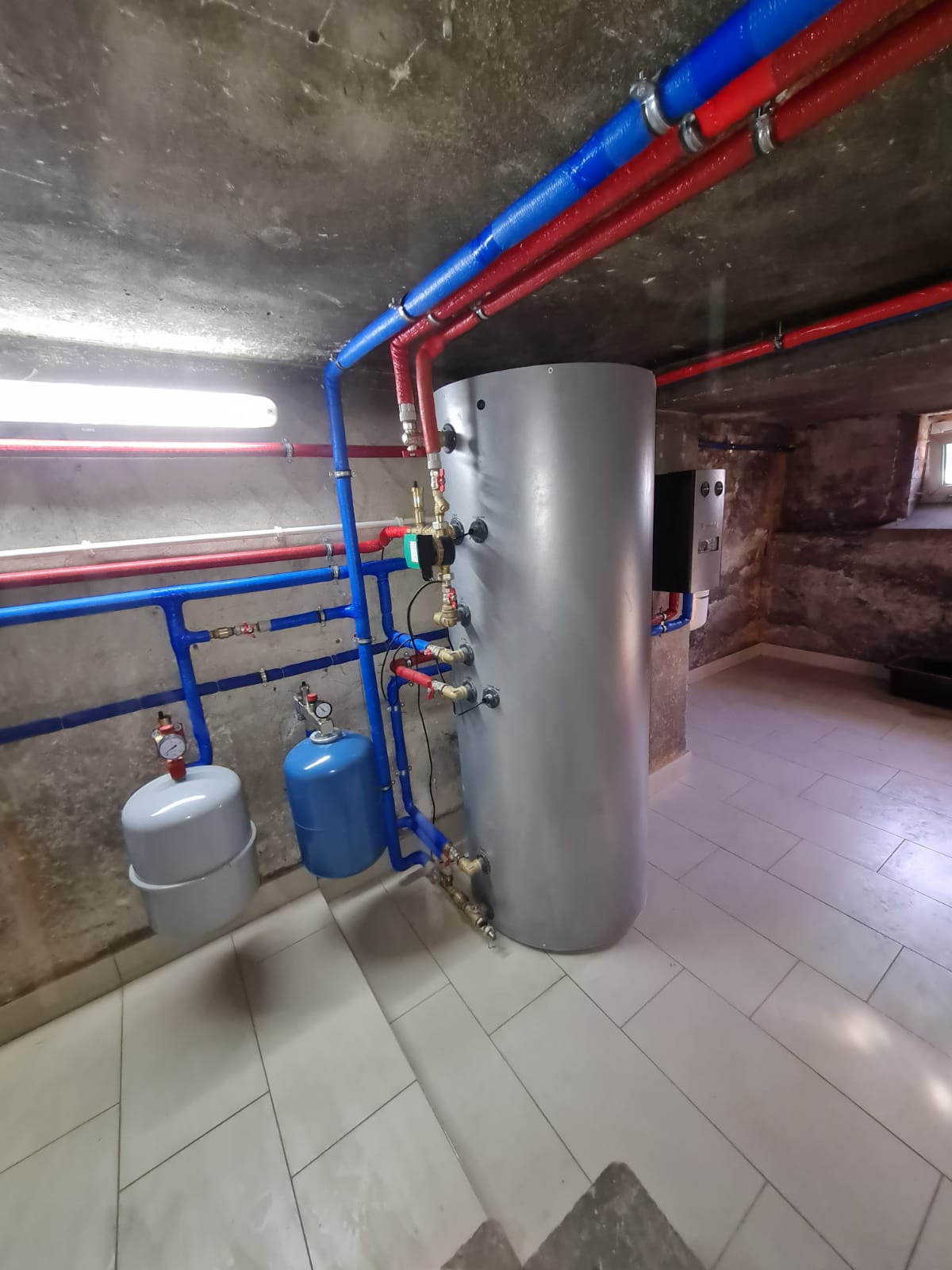 Szary zasobnik ciepłej wody użytkowej z niebieskimi i czerwonymi rurami instalacji grzewczej w piwnicy z surowym betonowym sufitem i płytkami na podłodze.