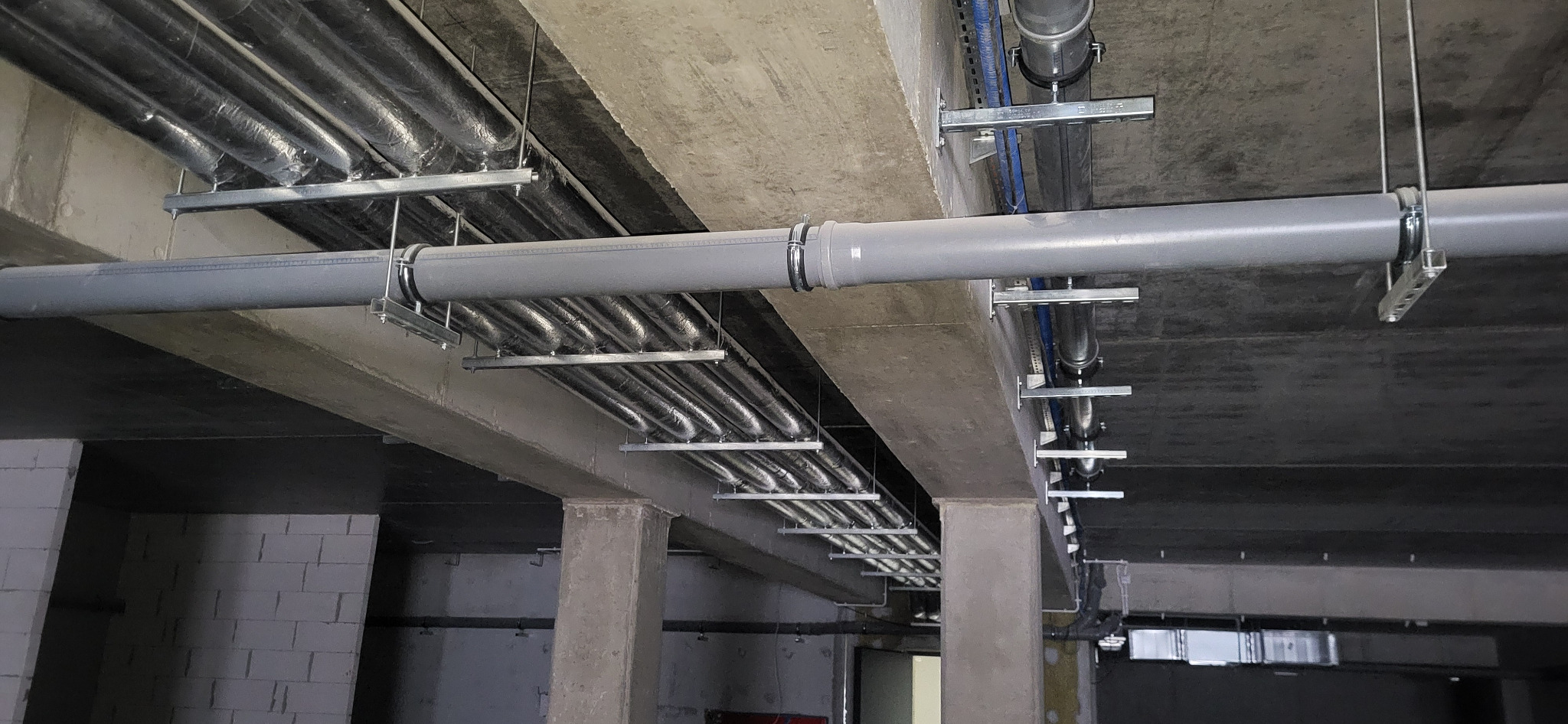Szare rury kanalizacyjne podwieszone na metalowych uchwytach pod betonowym sufitem w surowym stanie deweloperskim.