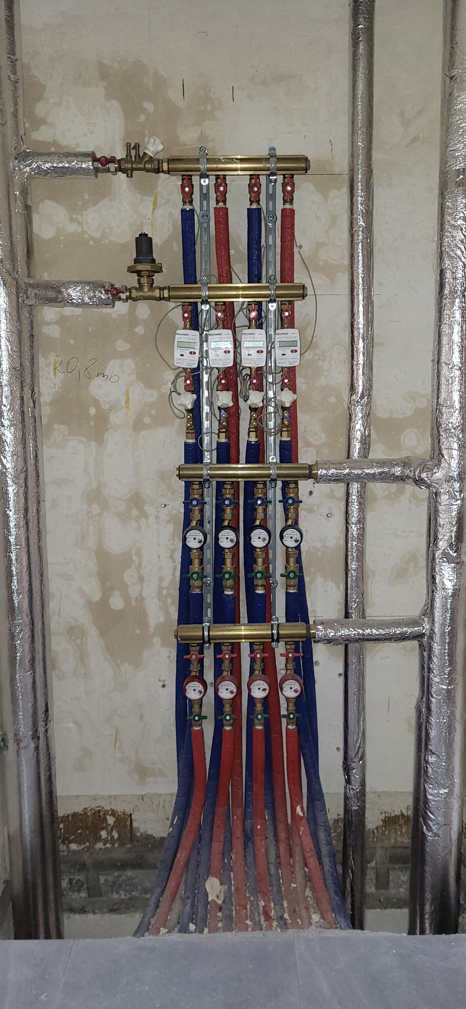 Rozdzielacz instalacji z rurami w izolacji czerwonej i niebieskiej, liczniki ciepła na pionach, rury owinięte folią aluminiową, surowa ściana w tle.