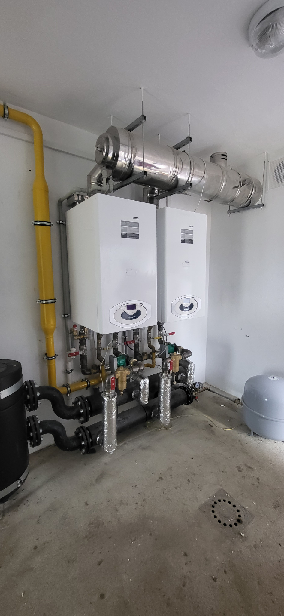 Dwa białe kotły grzewcze Ariston zamontowane na ścianie, z widocznymi rurami, zaworami, żółtą rurą gazową i srebrnymi przewodami wentylacyjnymi.