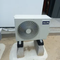 Zewnętrzna jednostka klimatyzacji marki AUX, zamontowana na betonowych bloczkach z amortyzującymi podkładkami, umieszczona na przygotowanym podłożu przed budynkiem.