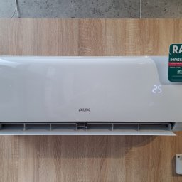Termserwis Jacek Sapak - Nowoczesny, biały klimatyzator AUX z wyświetlaczem 25°C, zamontowany na ścianie o drewnianym wzorze. Widoczna naklejka 'Raty' Credit Agricole. Pilot obok.