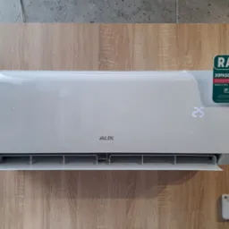 Nowoczesny, biały klimatyzator AUX z wyświetlaczem 25°C, zamontowany na ścianie o drewnianym wzorze. Widoczna naklejka 'Raty' Credit Agricole. Pilot obok.