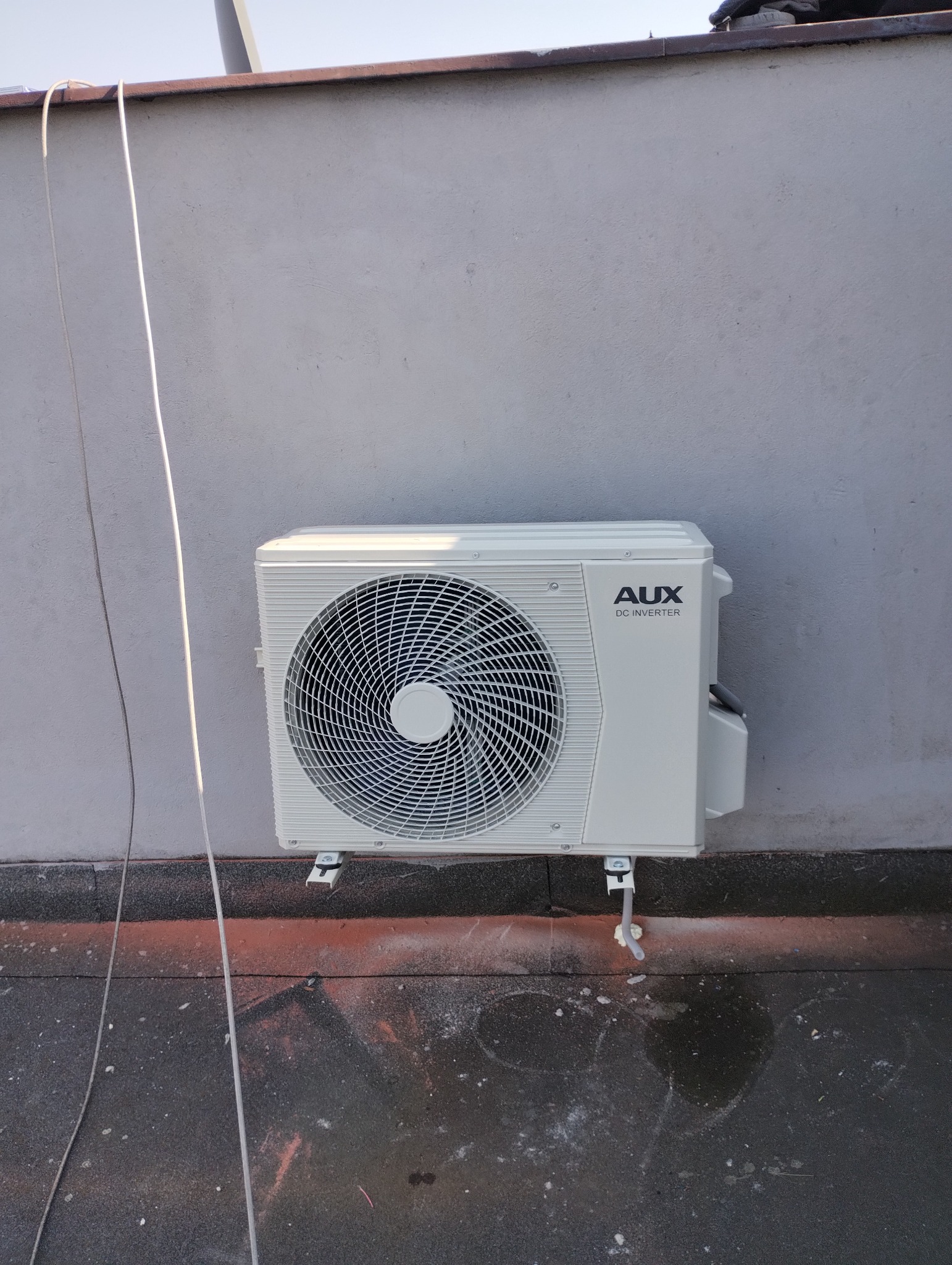Biała zewnętrzna jednostka klimatyzacji AUX DC Inverter zamontowana na szarej ścianie z widocznymi przewodami i elementami montażowymi na dachu pokrytym papą.