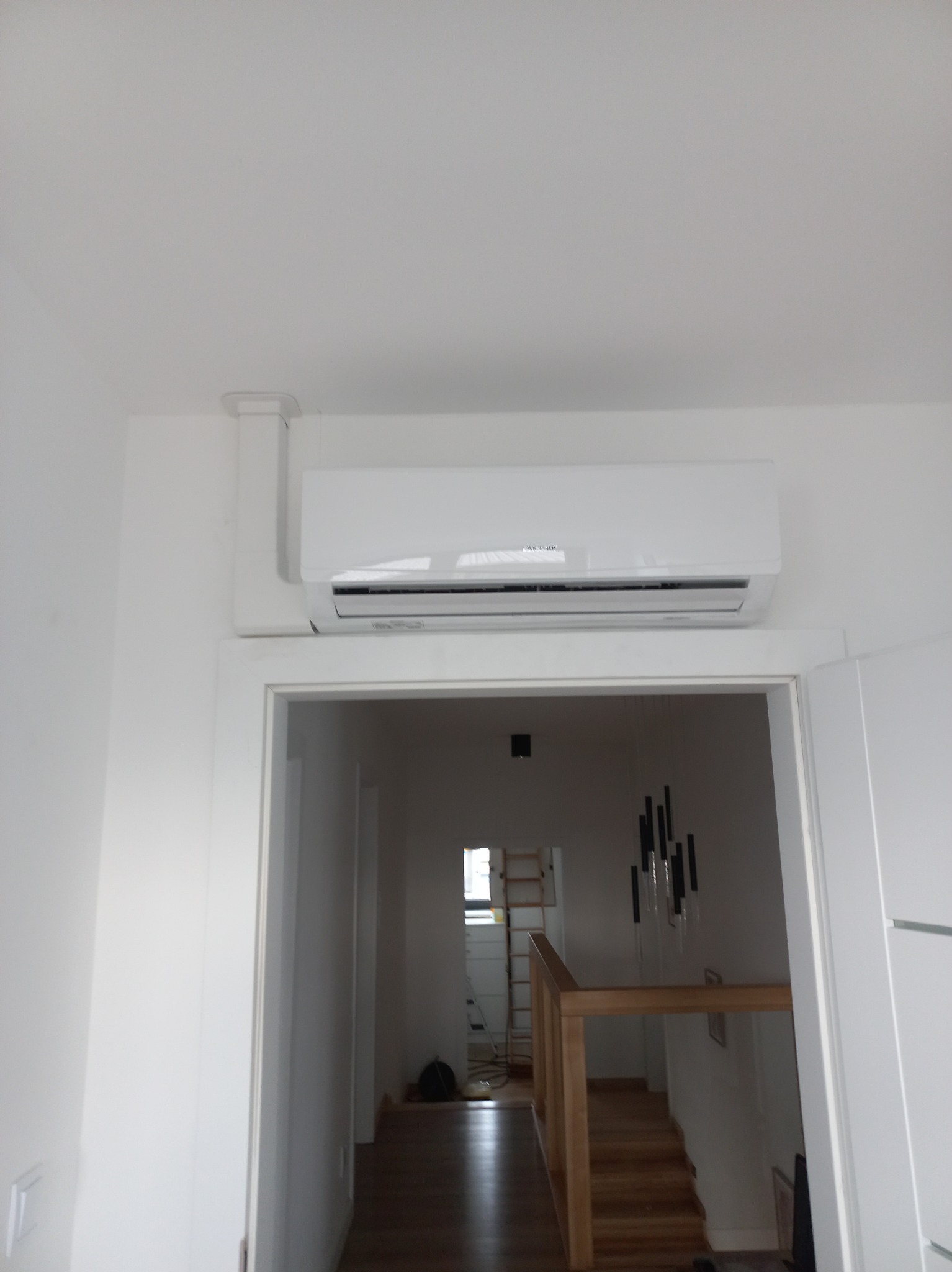 Biały klimatyzator zamontowany nad framugą drzwi w jasnym wnętrzu, widoczna instalacja odprowadzająca skropliny, w tle schody i drabina.