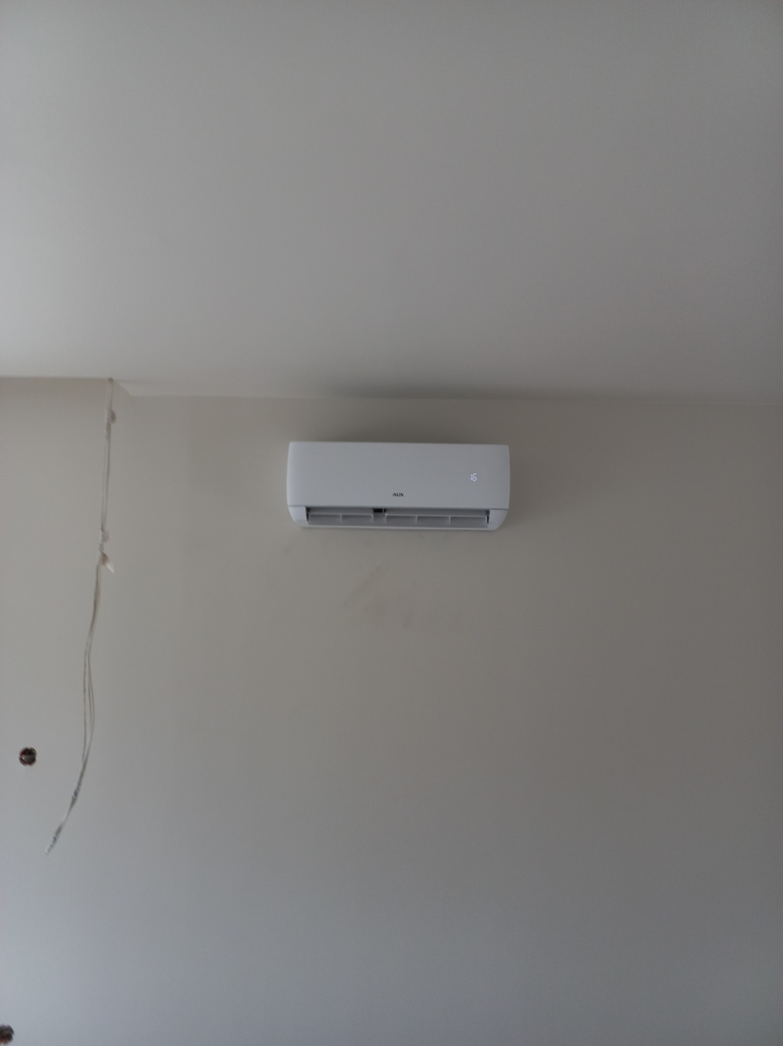 Biały klimatyzator marki AUX zamontowany na szarej ścianie w nowym pomieszczeniu, widoczne wystające przewody elektryczne i otwory po puszkach instalacyjnych.