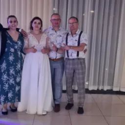 Grupa osób, w tym panna młoda w białej sukni, pozuje do zdjęcia w pomieszczeniu z białymi zasłonami i widocznym otoczeniem na zewnątrz, wszyscy pokazują kciuki w górę.