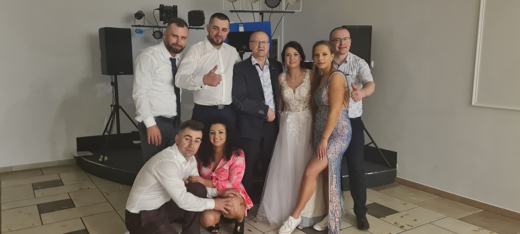 Grupa osób, w tym panna młoda w białej sukni z koronkową górą i goście w eleganckich strojach, pozuje na tle sprzętu nagłośnieniowego i oświetleniowego w sali weselnej.