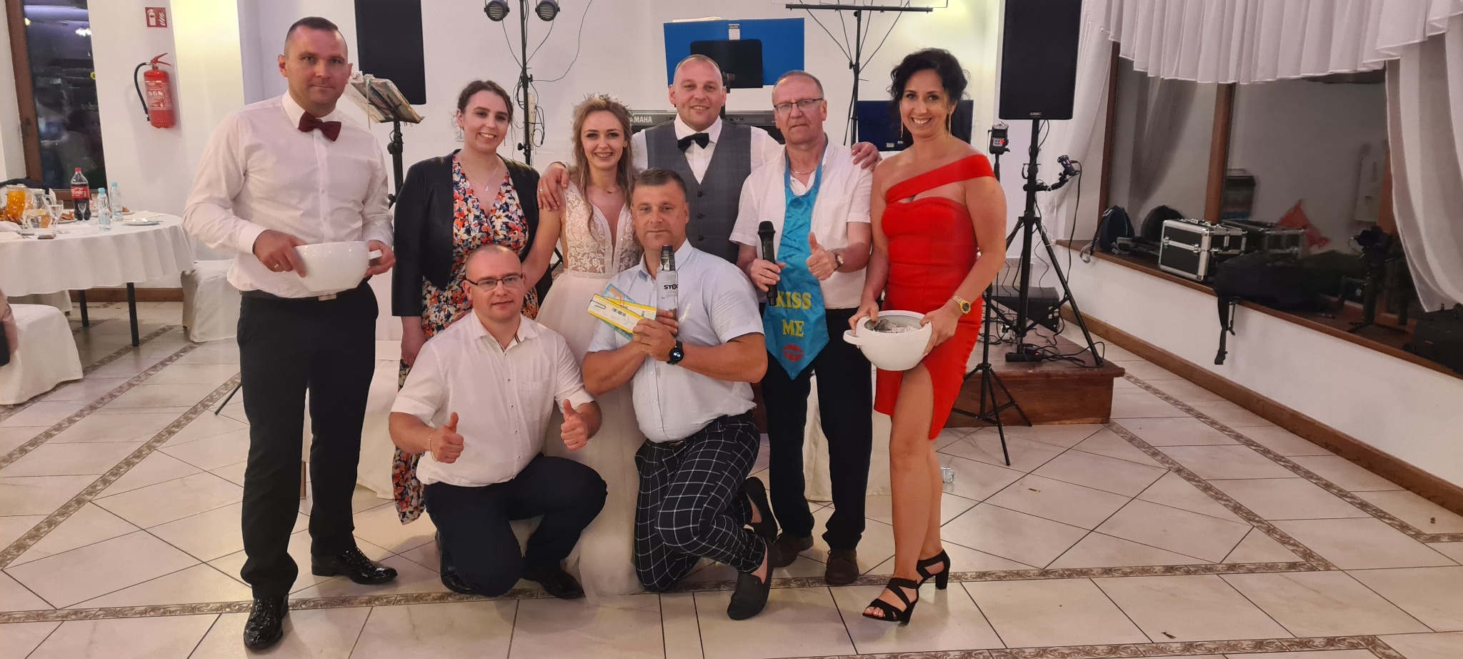 Grupa osób, w tym panna młoda, pozuje do zdjęcia z rekwizytami, takimi jak mikrofon, butelka wódki i miski na datki, prawdopodobnie podczas wesela.