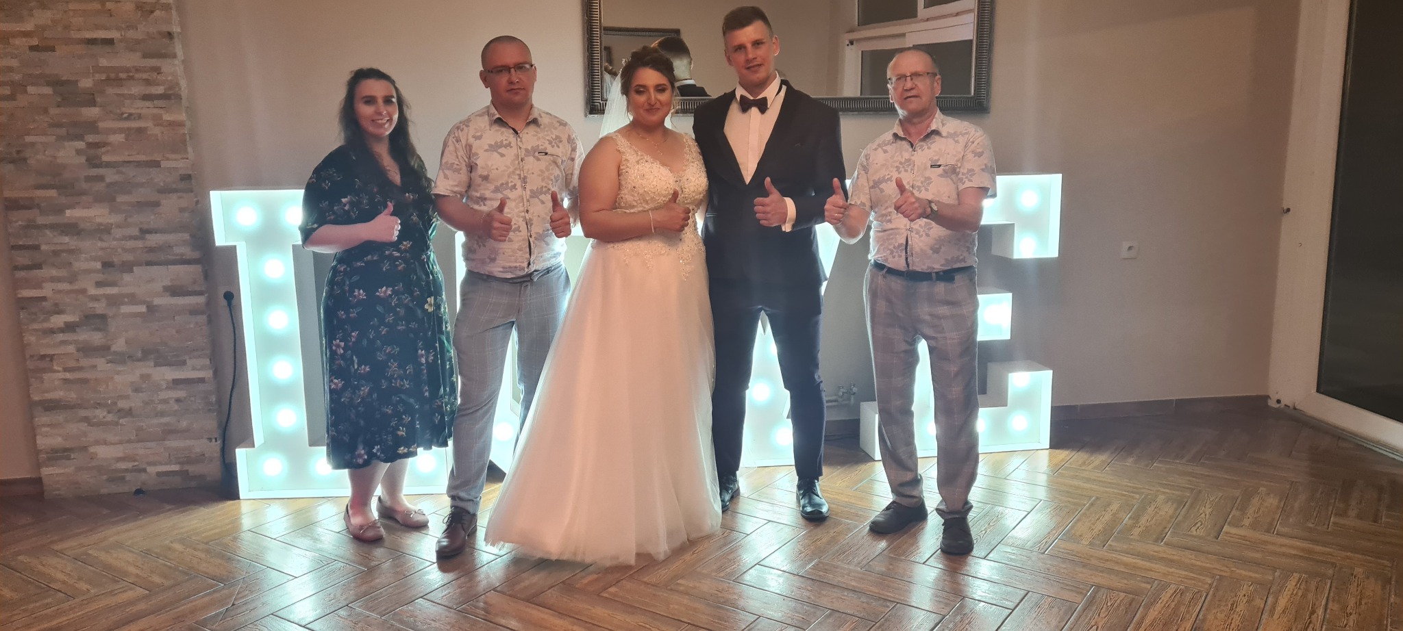 Pamiątkowe zdjęcie z wesela w Reszel, z panną młodą w białej sukni, panem młodym w smokingu i trzema gośćmi z uniesionymi kciukami na tle podświetlanych liter dekoracyjnych.
