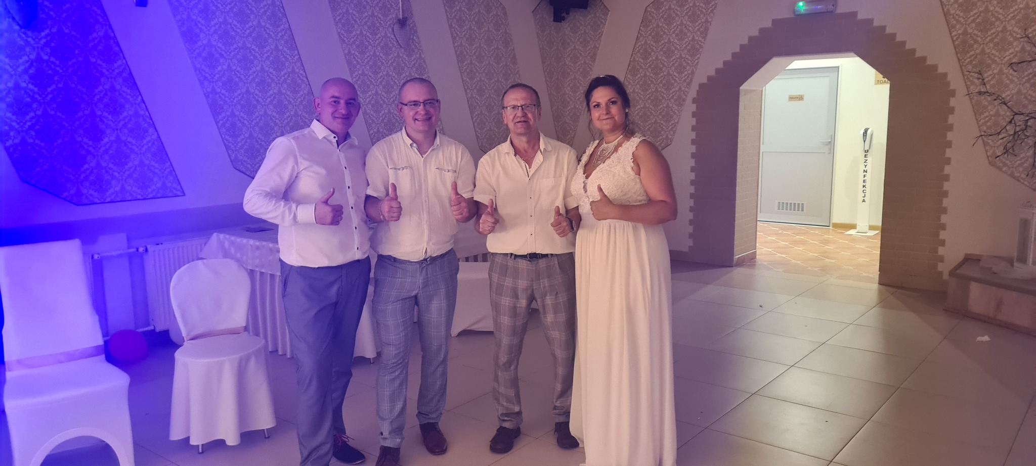 Grupa czterech osób, w tym kobieta w białej sukni, pozuje z uniesionymi kciukami w górę w sali z dekoracyjnymi ścianami i oświetleniem w odcieniach fioletu.