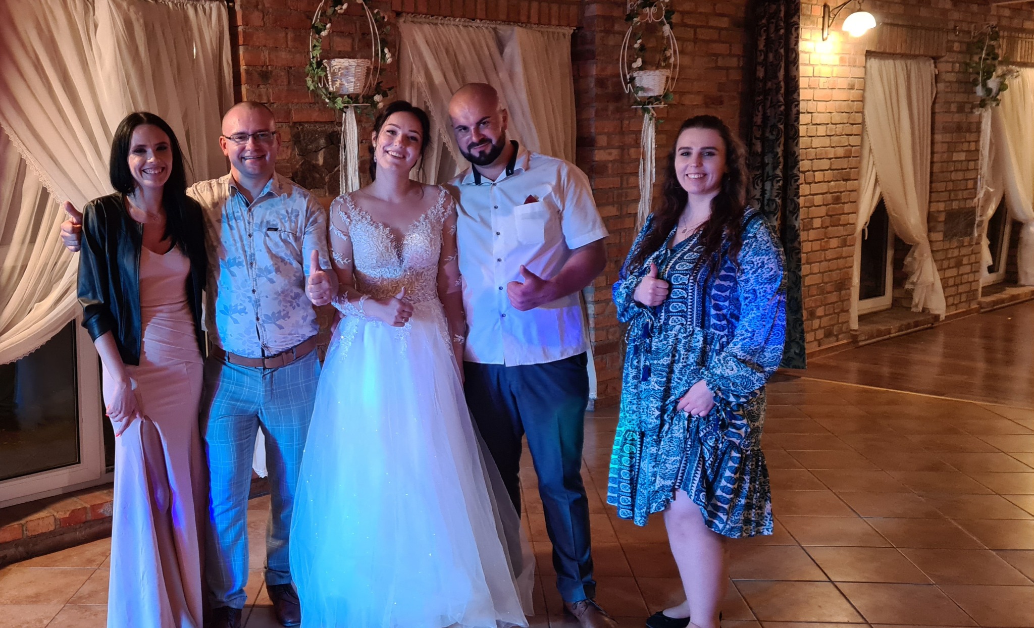 Pamiątkowe zdjęcie z wesela: para młoda w otoczeniu gości, wszyscy uśmiechnięci i z uniesionymi kciukami, ceglana ściana w tle.