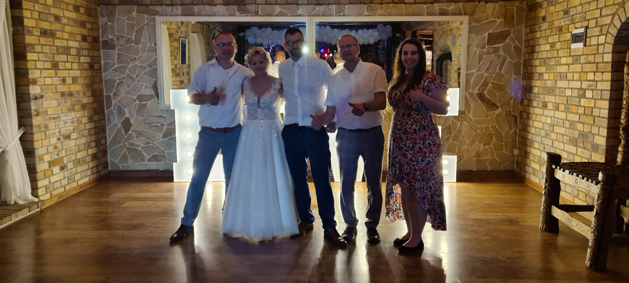 Zdjęcie grupy osób, w tym panna młoda w białej sukni, pozujących na tle dekoracji z podświetlanych liter w sali z kamienną ścianą.