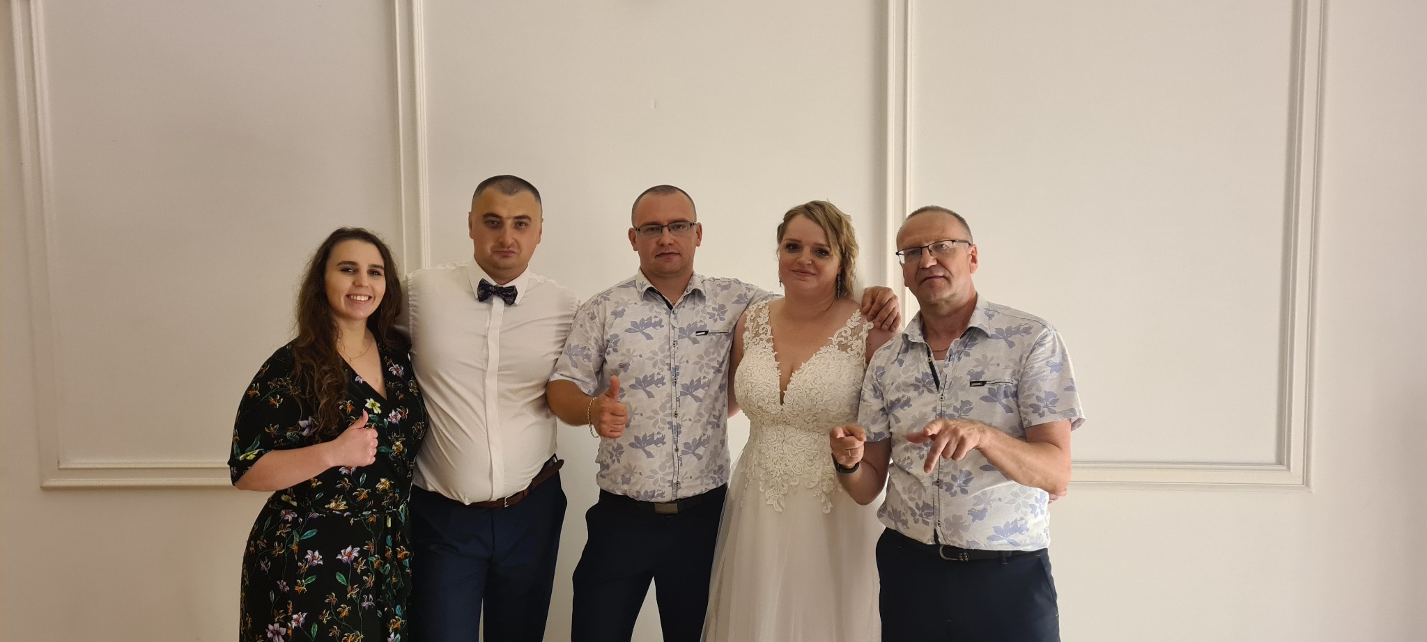 Grupa pięciu osób, w tym panna młoda w koronkowej sukni, pozuje na jasnym tle z dekoracyjnymi ramkami, mężczyźni w koszulach z krótkim rękawem i muszką, kobiety w kwiecistych sukienkach, wszyscy...