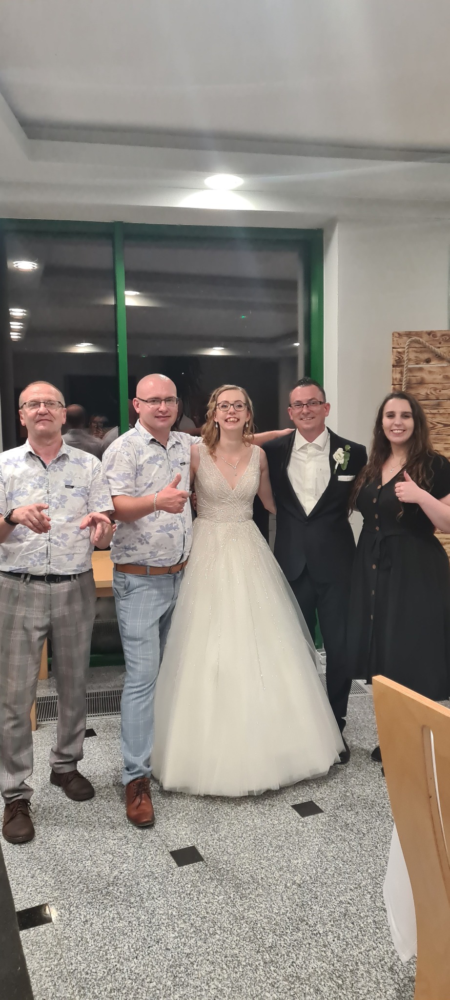 Panna młoda w sukni ślubnej pozuje z gośćmi weselnymi, mężczyźni ubrani w koszule w kwiaty i spodnie w kratę, kobieta w czarnej sukience, wszyscy uśmiechnięci i z podniesionymi kciukami.