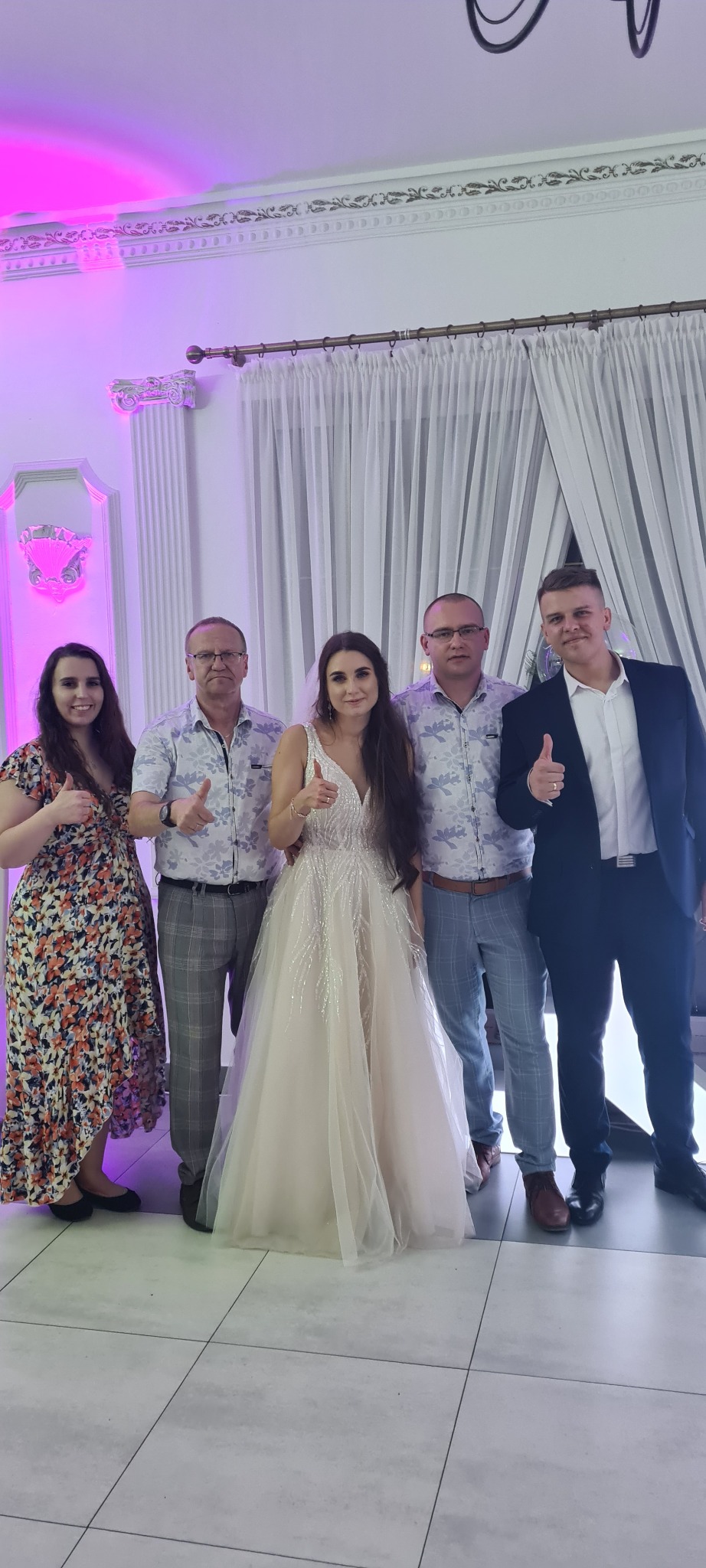 Panna młoda w białej sukni ślubnej pozuje z gośćmi, wszyscy pokazują kciuki w górę na tle sali z kolumnami i białymi zasłonami.