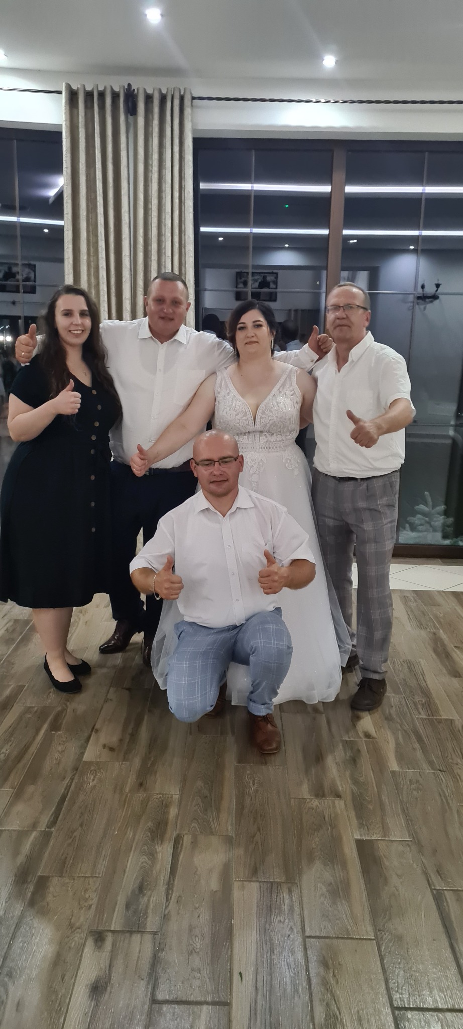 Panna młoda w białej sukni ślubnej pozuje do zdjęcia z gośćmi weselnymi, wszyscy z uniesionymi kciukami w górę, na drewnianej podłodze sali weselnej z dużymi oknami.