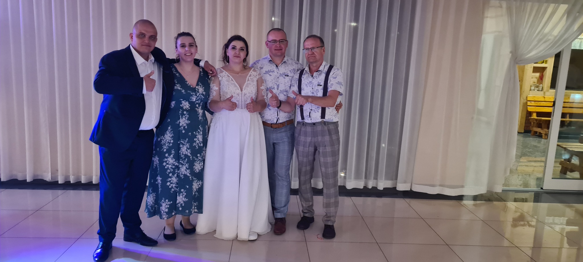 Grupa osób, w tym panna młoda w białej sukni, pozuje do zdjęcia w pomieszczeniu z białymi zasłonami i widocznym otoczeniem na zewnątrz, wszyscy pokazują kciuki w górę.