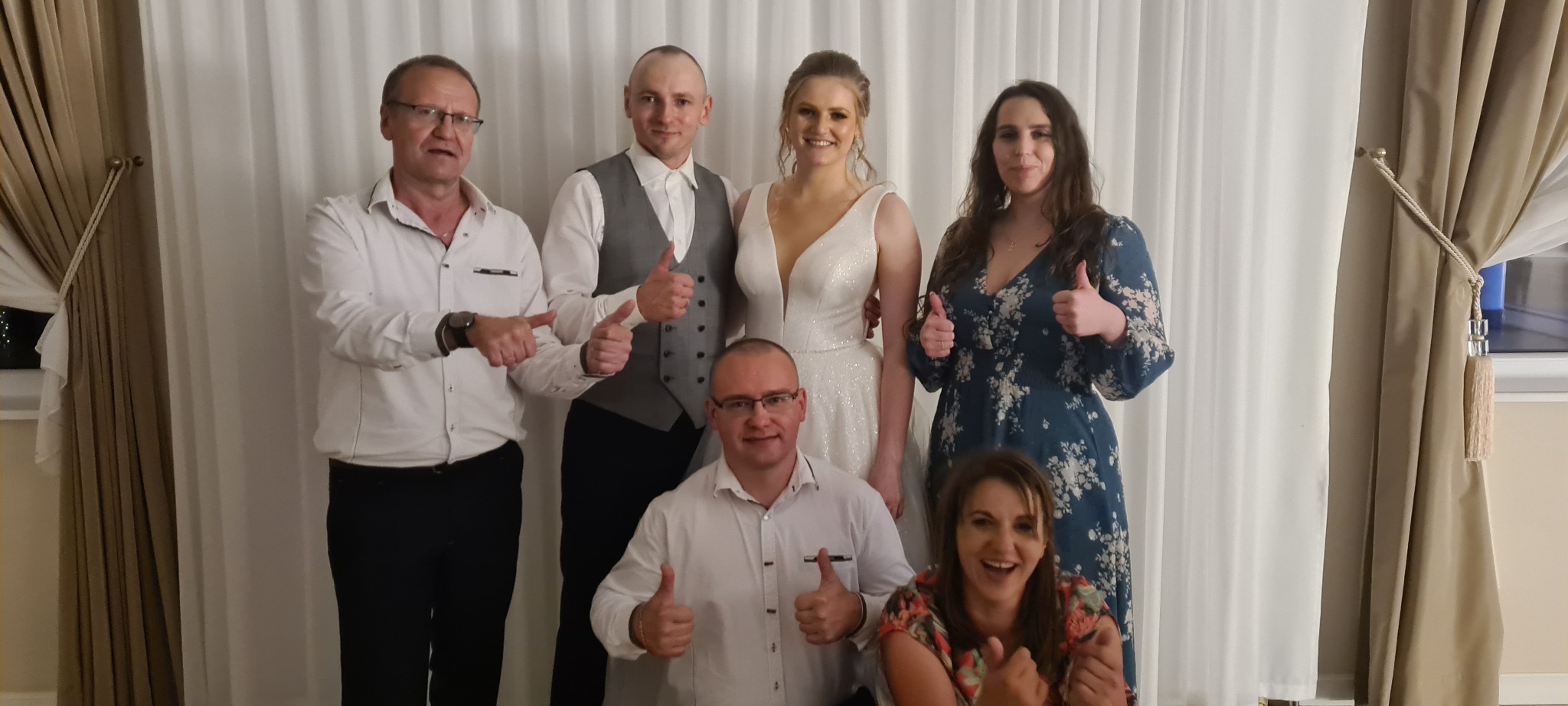 Zespół muzyczny z panną młodą i gośćmi weselnymi pozuje do zdjęcia, wszyscy pokazują kciuki w górę na tle białej zasłony.