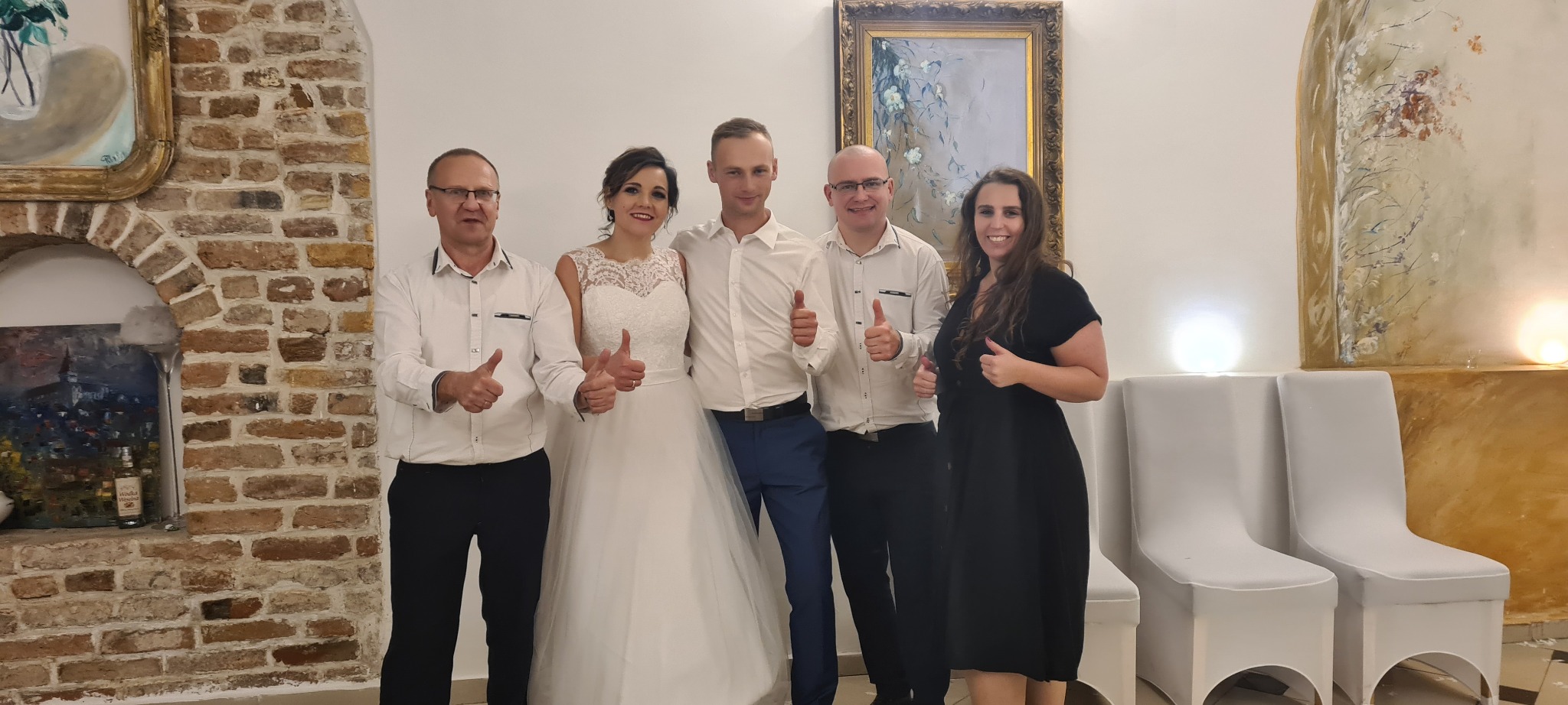 Grupa osób, w tym para młoda w sukni ślubnej, pozuje do zdjęcia z uniesionymi kciukami w pomieszczeniu z ceglaną ścianą i obrazami.