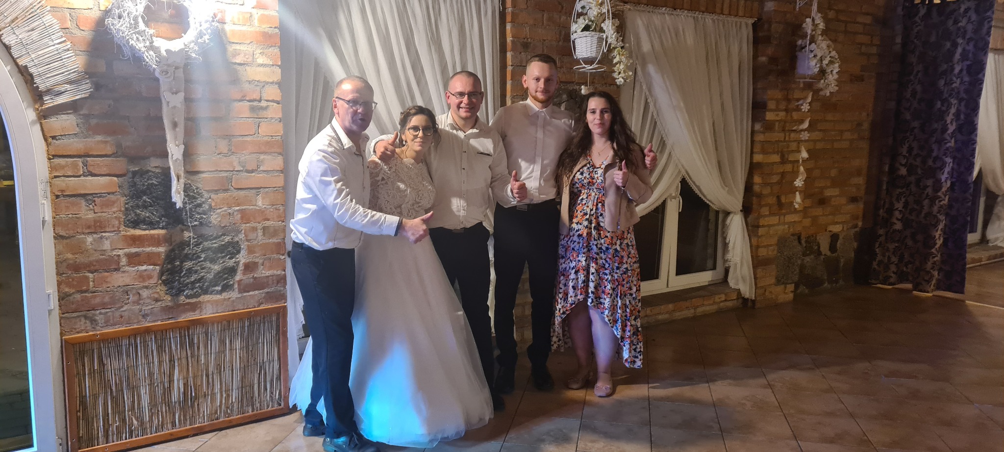 Zdjęcie z wesela: Panna młoda w koronkowej sukni pozuje z trzema mężczyznami i kobietą, wszyscy z uniesionymi kciukami na tle ceglanej ściany z dekoracjami i zasłonami.