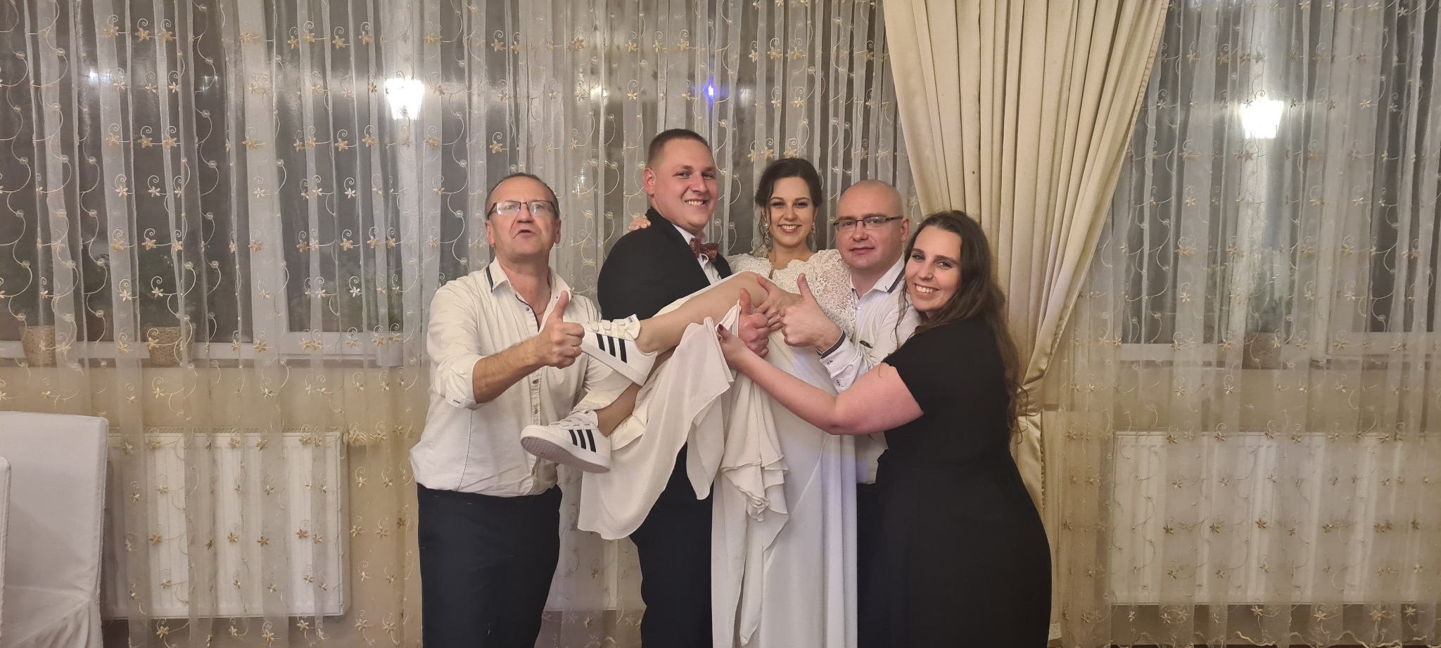 Uśmiechnięta panna młoda w białej sukni i sportowych butach Adidas, trzymana przez pana młodego w towarzystwie gości weselnych, wszyscy z uniesionymi kciukami na tle dekoracyjnych firan.