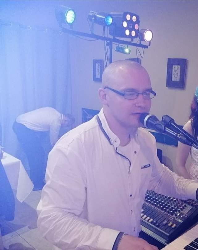 Mężczyzna w okularach śpiewa do mikrofonu, stojąc za keyboardem na tle oświetlenia scenicznego i innych osób.