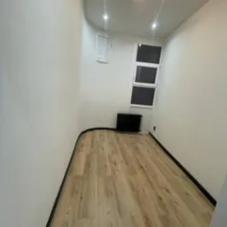 Jasne panele podłogowe ułożone w małym, odświeżonym pokoju z białymi ścianami i czarnymi listwami przypodłogowymi, oświetlonym nowoczesną lampą sufitową.