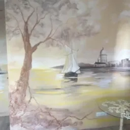 Iluzjonistyczne malowidło ścienne przedstawiające pejzaż morski z latarnią morską, żaglówką i architekturą miejską w sepii, z elementami drzewa wkomponowanymi w narożnik pokoju.