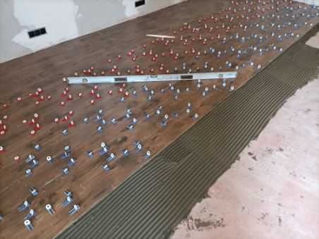 Proces układania drewnopodobnych płytek z użyciem systemu poziomowania: poziomica, kliny i klipsy widoczne na powierzchni pokrytej klejem.