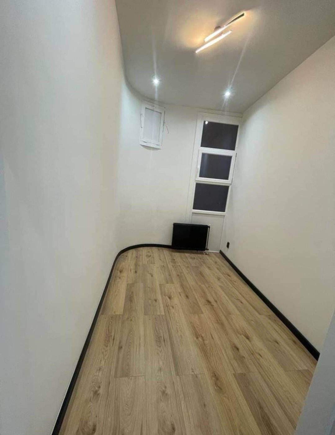 Wąskie pomieszczenie z ułożonymi panelami podłogowymi w odcieniach beżu i brązu, białe ściany, widoczne czarne listwy przypodłogowe i grzejnik, oświetlenie sufitowe.