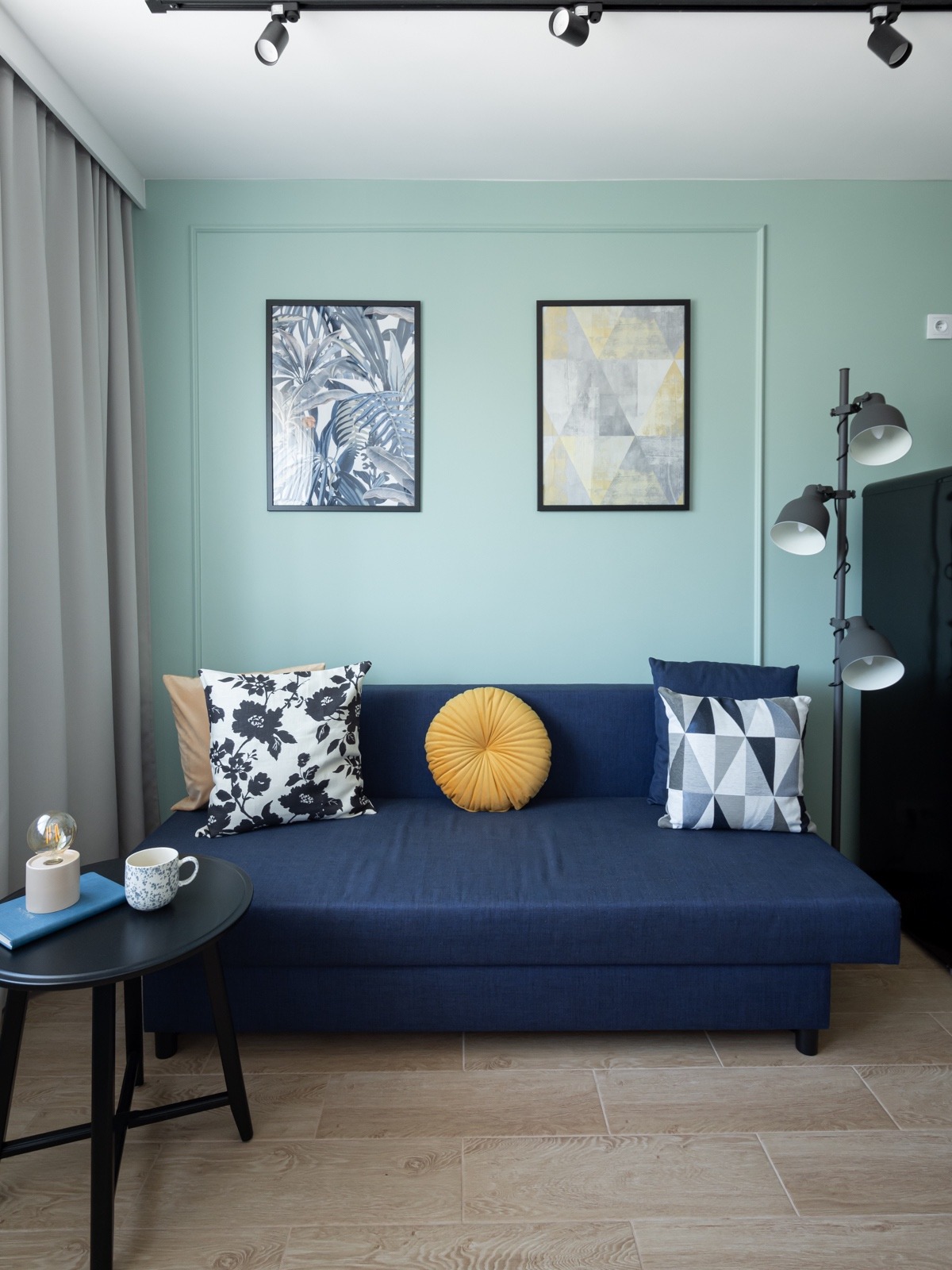 Niebieska sofa z dekoracyjnymi poduszkami i okrągłą, żółtą poduszką, ustawiona na drewnopodobnych panelach, w tle miętowa ściana z dwoma obrazami w czarnych ramkach, oświetlenie szynowe na suficie...