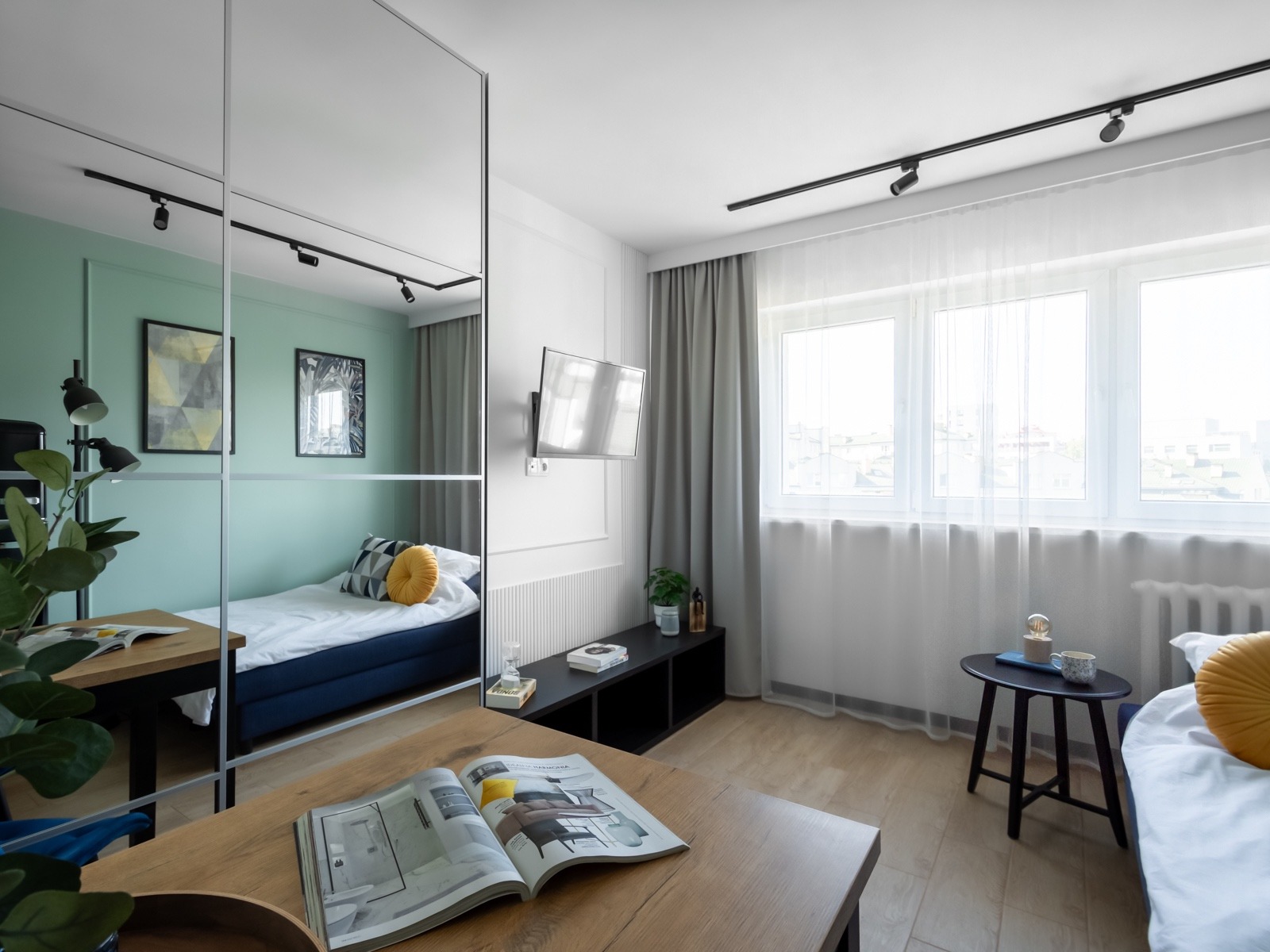 Jasne, nowocześnie urządzone wnętrze pokoju dziennego z łóżkiem widocznym w lustrzanej szafie, drewniany stolik kawowy z otwartym magazynem o wnętrzach, okno z białymi firanami.