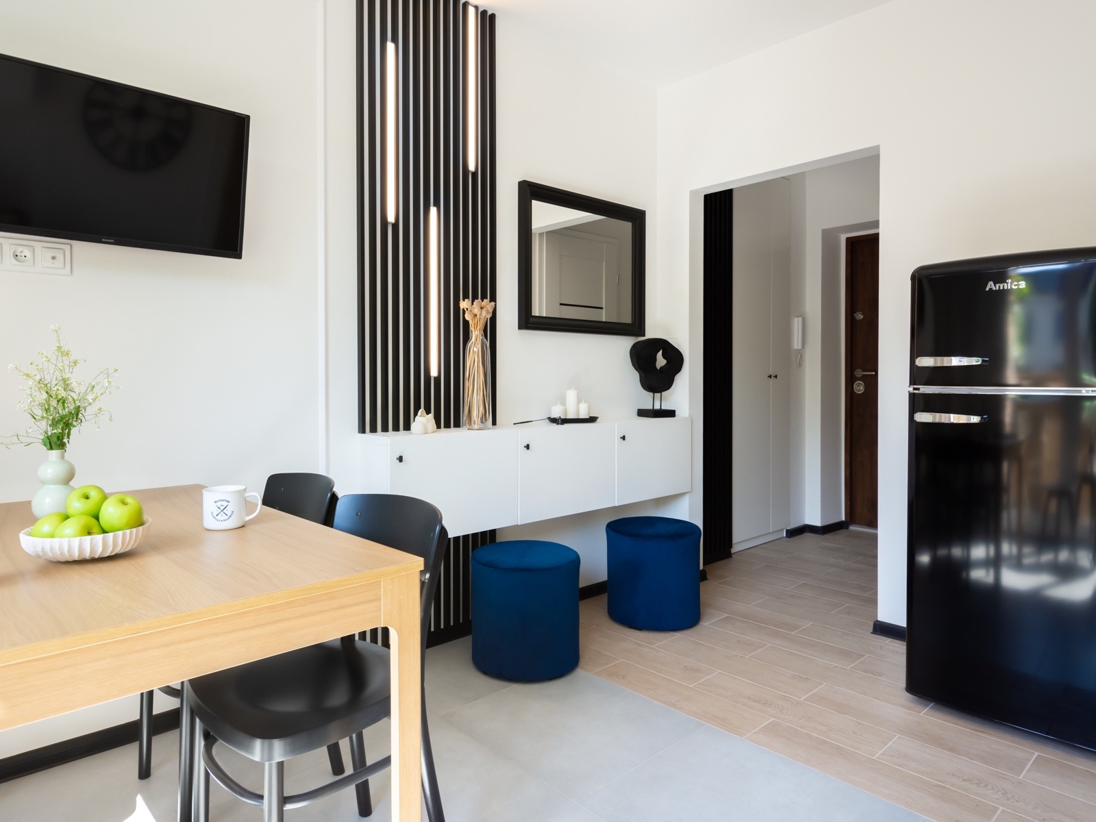Jasne wnętrze mieszkania z drewnianym stołem, czarnymi krzesłami, białym kredensem z dekoracjami, niebieskimi pufami i czarną lodówką w stylu retro.