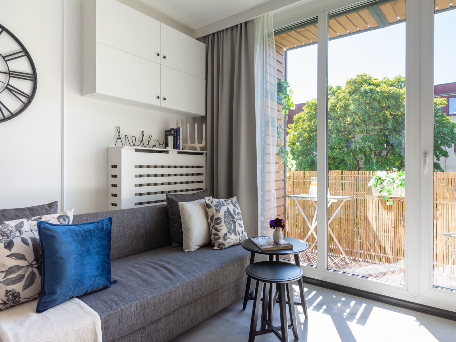 Jasne wnętrze salonu z szarą sofą, dekoracyjnymi poduszkami, białymi szafkami wiszącymi i widokiem na balkon z bambusową osłoną i stolikiem